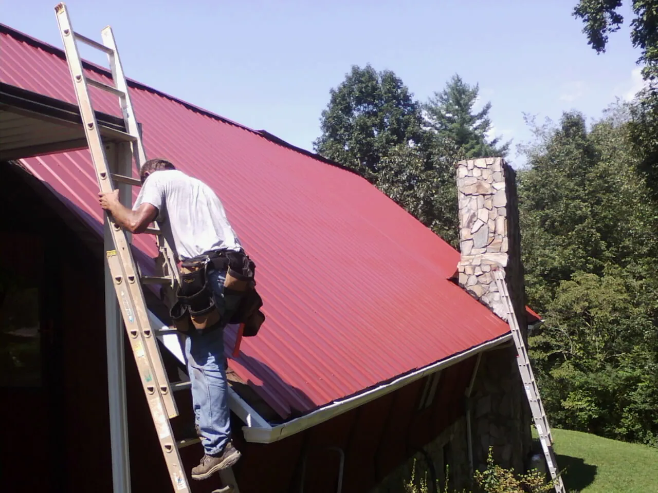 Roofer on a ladder beside a red metal roof and stone chimney — Soffit & Fascia Repair by Roofing Brothers in Monee